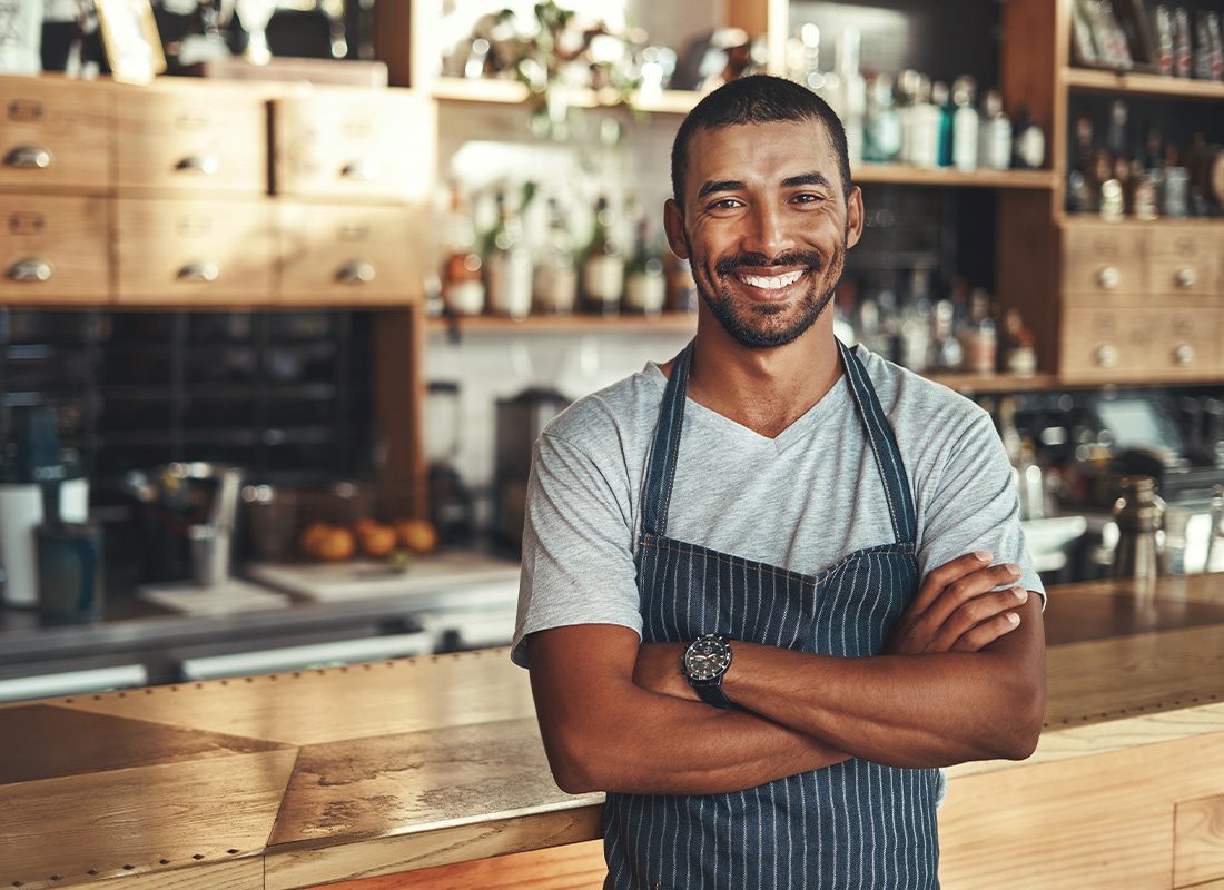 Business Insurance - Friendly Confident Business Owner Standing With Arms Crossed and Smiling in Front of the Counter at His Cafe